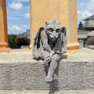 Seated Gargoyle with his legs hanging over the edge.  He is outside on cement with two orange columns in the background

House Parts home decor handmade Atlanta gargoyle grotesque whimsical fantasy mystical creature. Unique gift for fantasy fans, dungeon, or dinosaurs. Unique Christmas gift for someone who has everything. Gothic, medieval, unique, artisan, craft, detail, specialty, a gift that no one else has, special, protective, family small business, local, odd, quirky gift