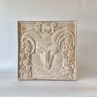 Decorative wall art relief of a ram's head with fruit and flowers surrounding the ram's face on a white background